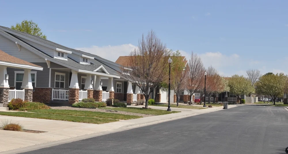 Residential street with modern single-story houses, neatly lined trees, and a clear sky. The road is empty and well-paved. MHPI Base Housing Rights